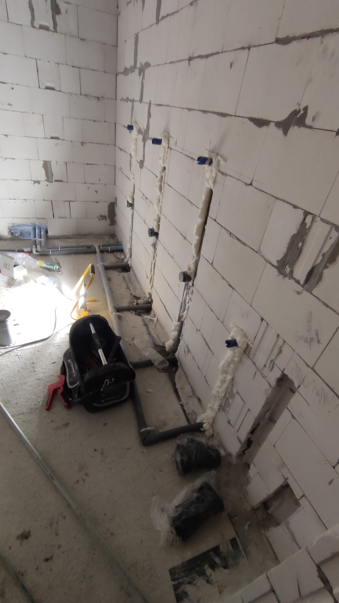 Ściana z betonu komórkowego z wyciętymi bruzdami wypełnionymi pianką montażową wokół rur instalacji wodnej, torba narzędziowa leżąca na podłodze.