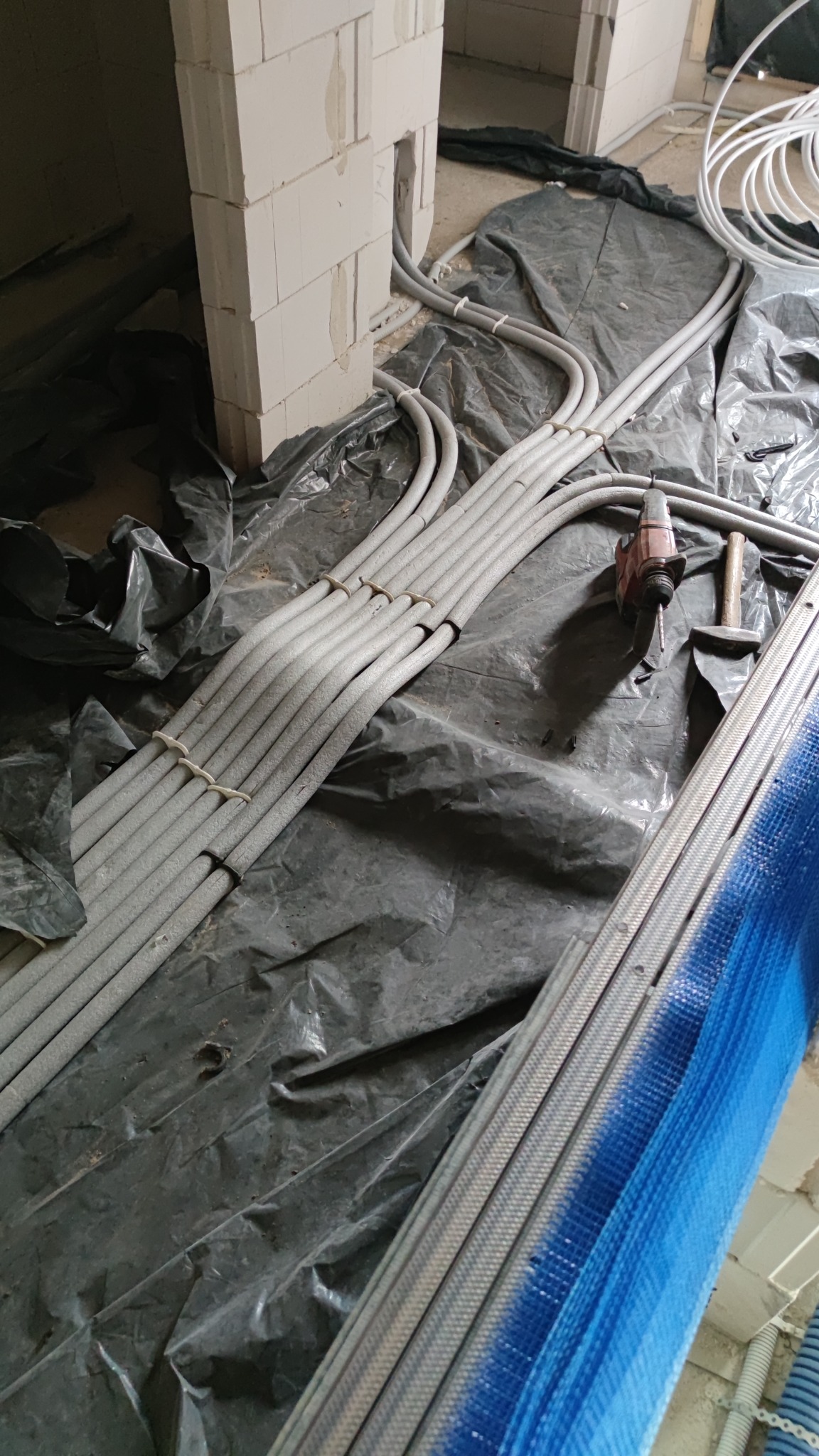 Szare rury instalacyjne ułożone na folii budowlanej, przymocowane opaskami, obok wiertarka i młotek, widok na surowy mur z pustaków.