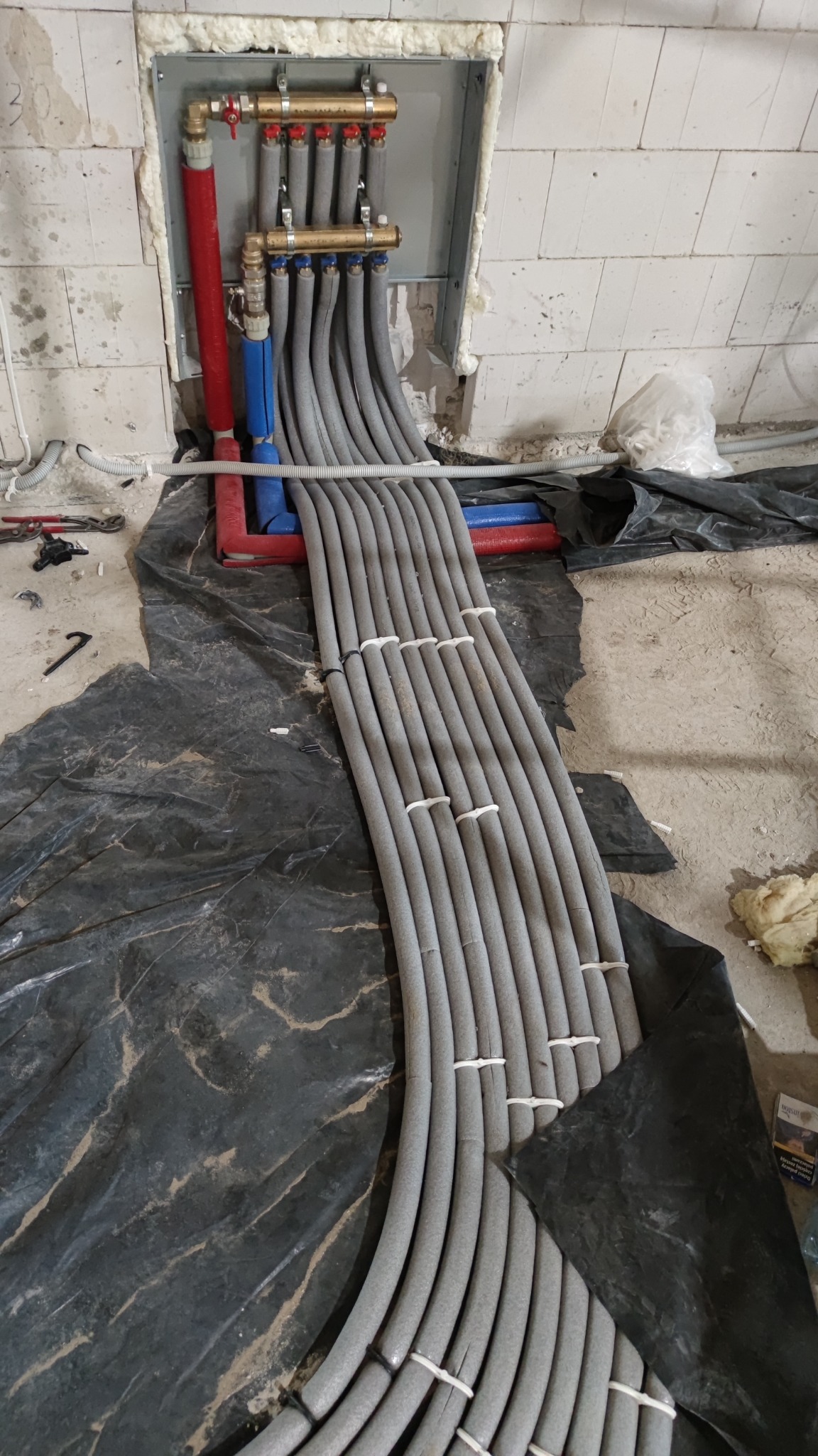 Rozdzielacz ogrzewania podłogowego z szarymi rurami w izolacji, częściowo ułożonymi na czarnej folii ochronnej, widoczne elementy instalacji w ścianie.