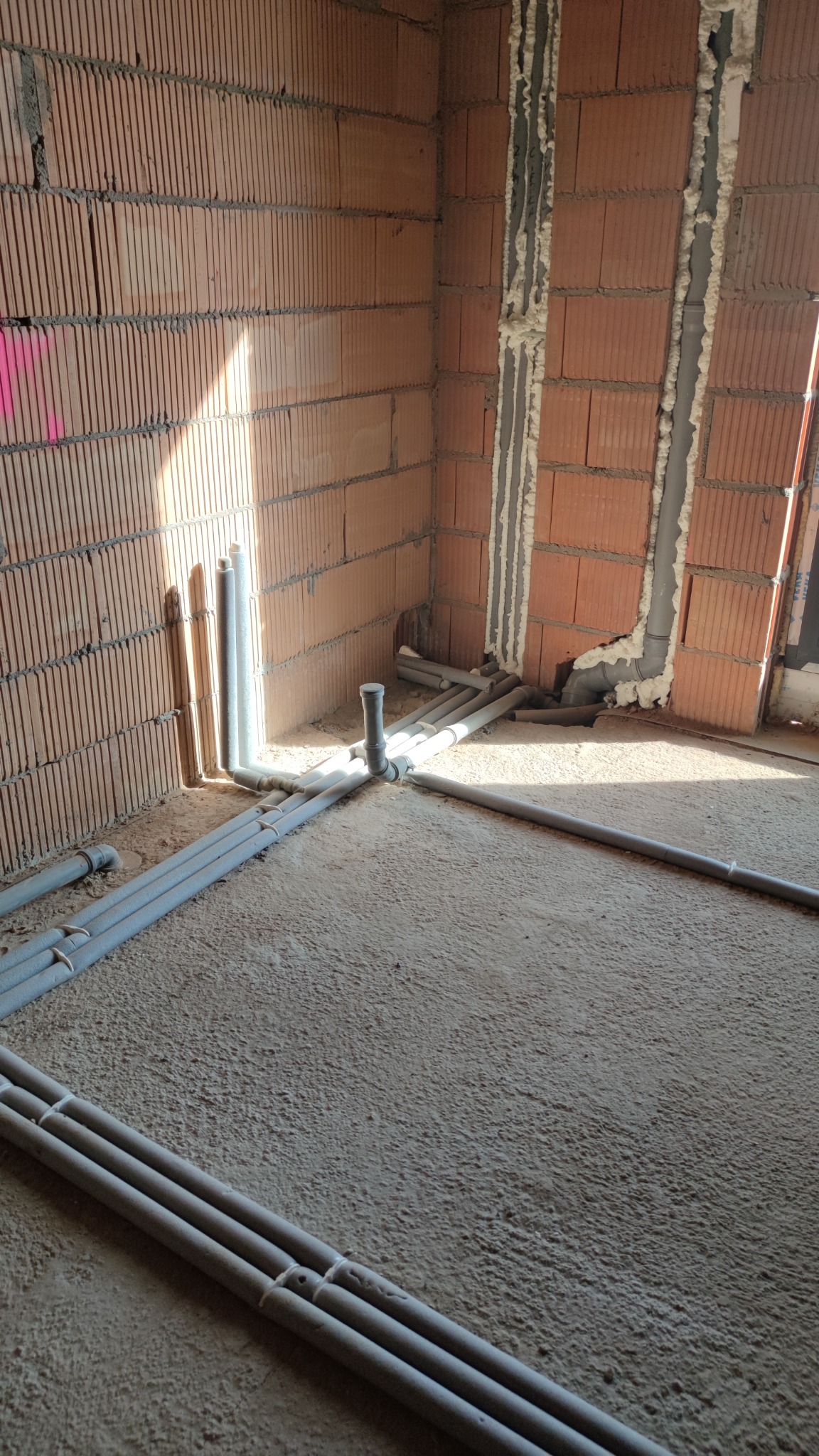 Surowe wnętrze z ceglanymi ścianami i szarymi rurami instalacji wodnej lub grzewczej ułożonymi na podłodze w trakcie montażu.