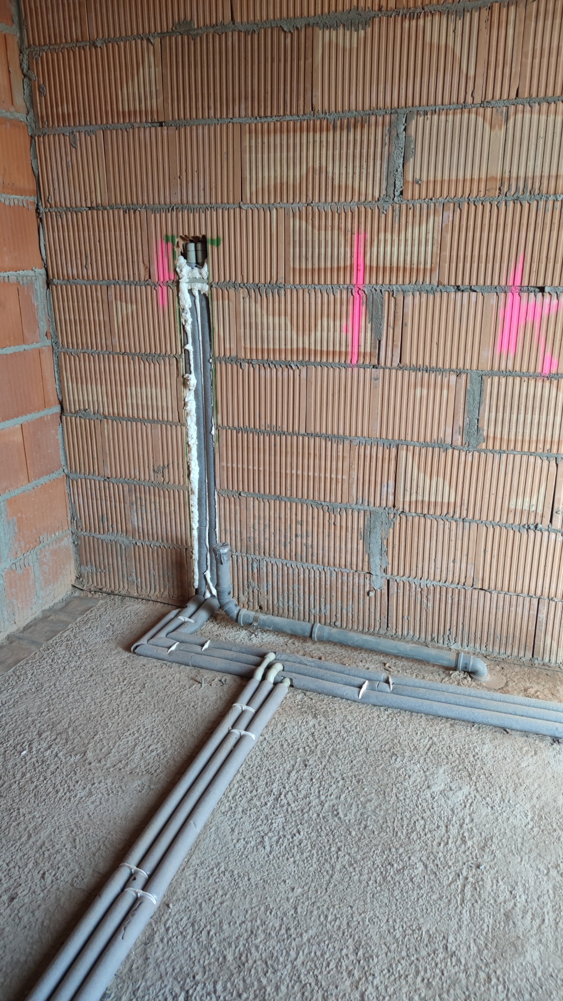 Surowy mur z cegły z zaznaczonymi różowymi liniami, pionowe rury instalacyjne uszczelnione pianką montażową, poziome szare rury na szarej, betonowej podłodze w trakcie instalacji.