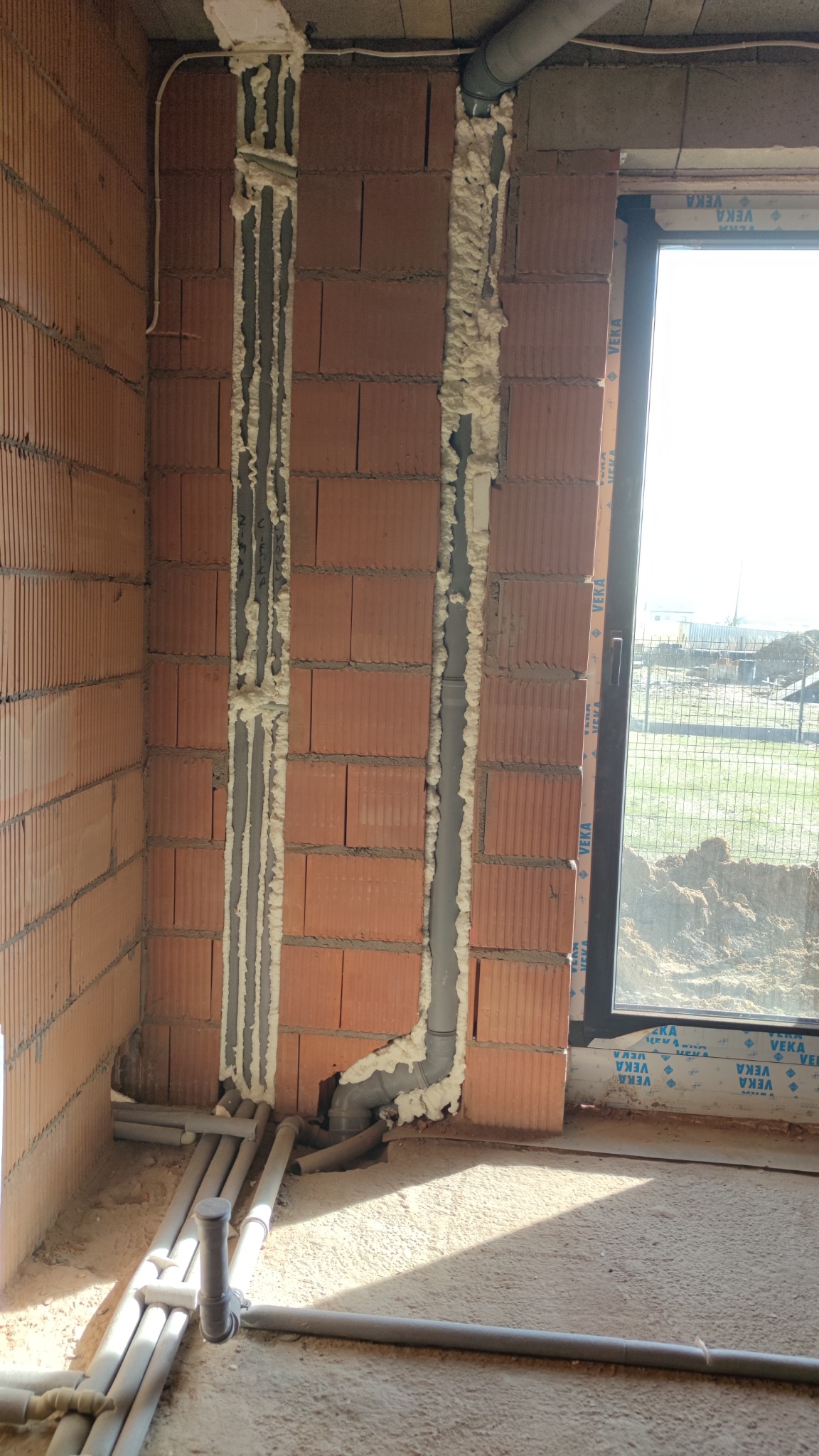 Wewnętrzne ściany z cegły z bruzdami wypełnionymi pianką montażową wokół szarych rur kanalizacyjnych, widoczne rury na podłodze w surowym stanie, okno z profilem VEKA.