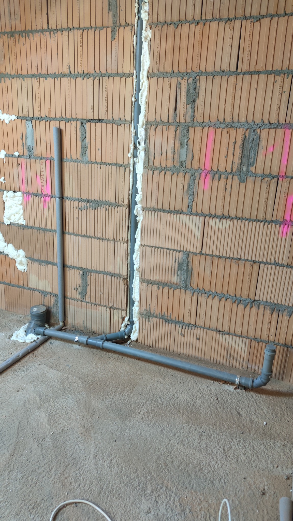 Szare rury kanalizacyjne z tworzywa sztucznego prowadzone wzdłuż ceglanej ściany z widoczną pianką montażową i oznaczeniami w kolorze różowym.