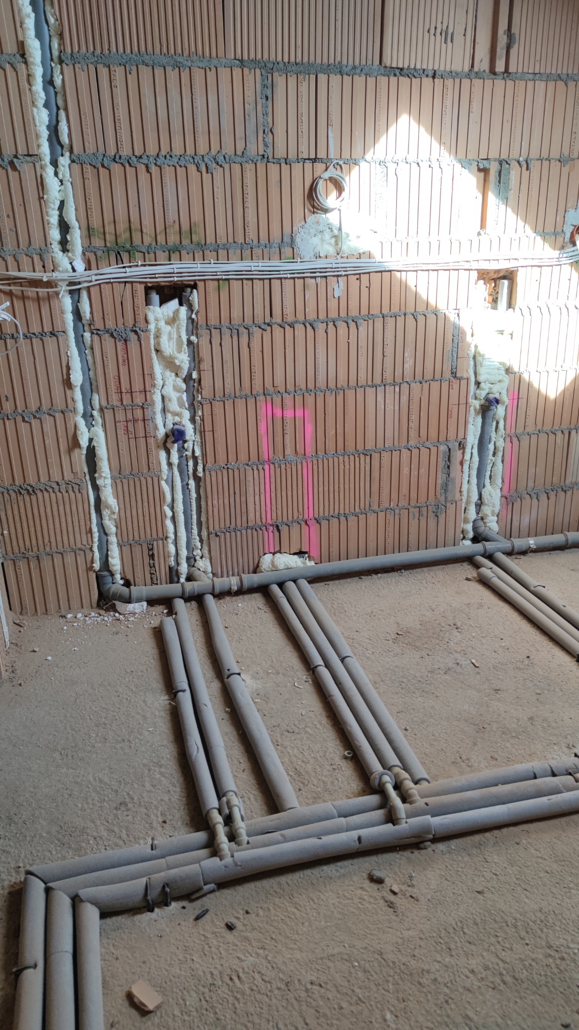 Surowa instalacja ogrzewania podłogowego w trakcie montażu: szare rury ułożone na posadzce, ściany z czerwonej cegły z pianką montażową wokół pionowych rur.