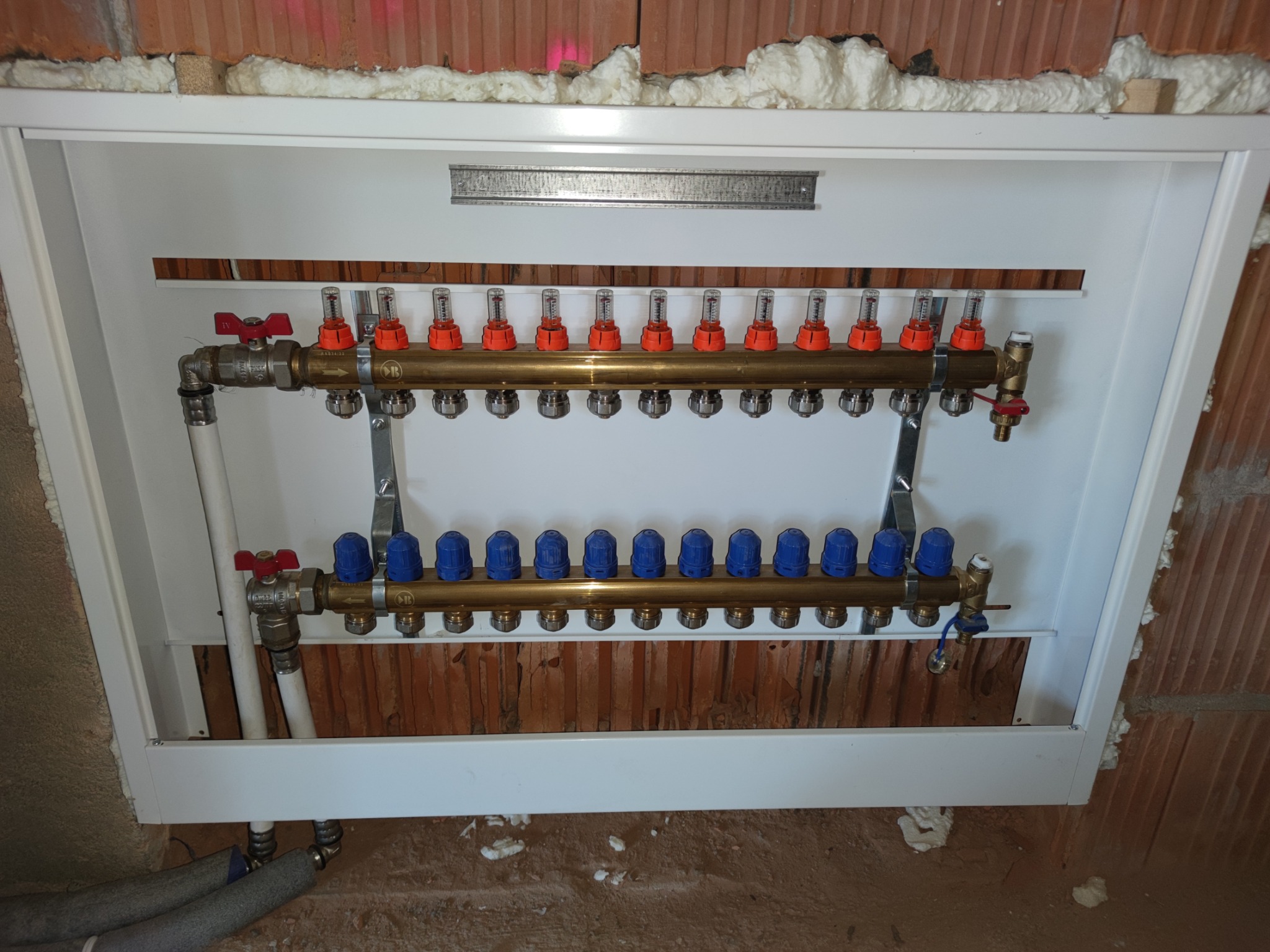Rozdzielacz ogrzewania podłogowego z czerwonymi i niebieskimi zaworami, zamontowany w białej szafce w surowym stanie wykończenia ściany z cegły i pianki montażowej.