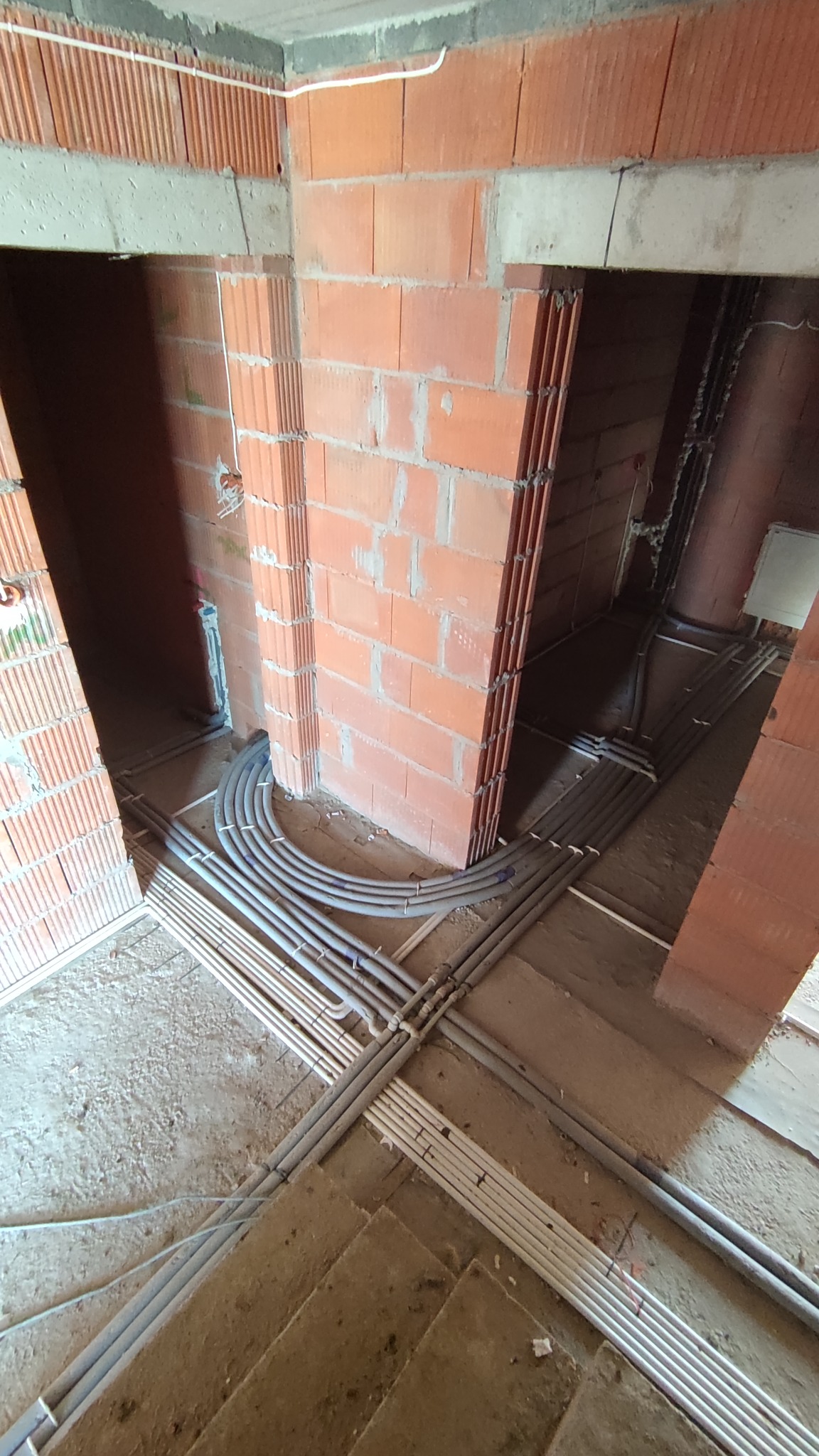 Surowy stan budynku w trakcie instalacji elektrycznej: widok na podłogę z licznymi rurkami instalacyjnymi prowadzącymi do ścian z czerwonej cegły.