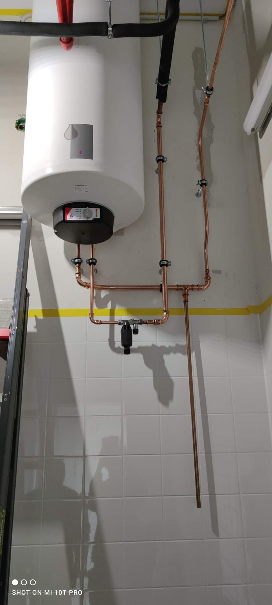 Biały bojler z miedzianymi rurami instalacji grzewczej na tle białych płytek i żółtej taśmy malarskiej.