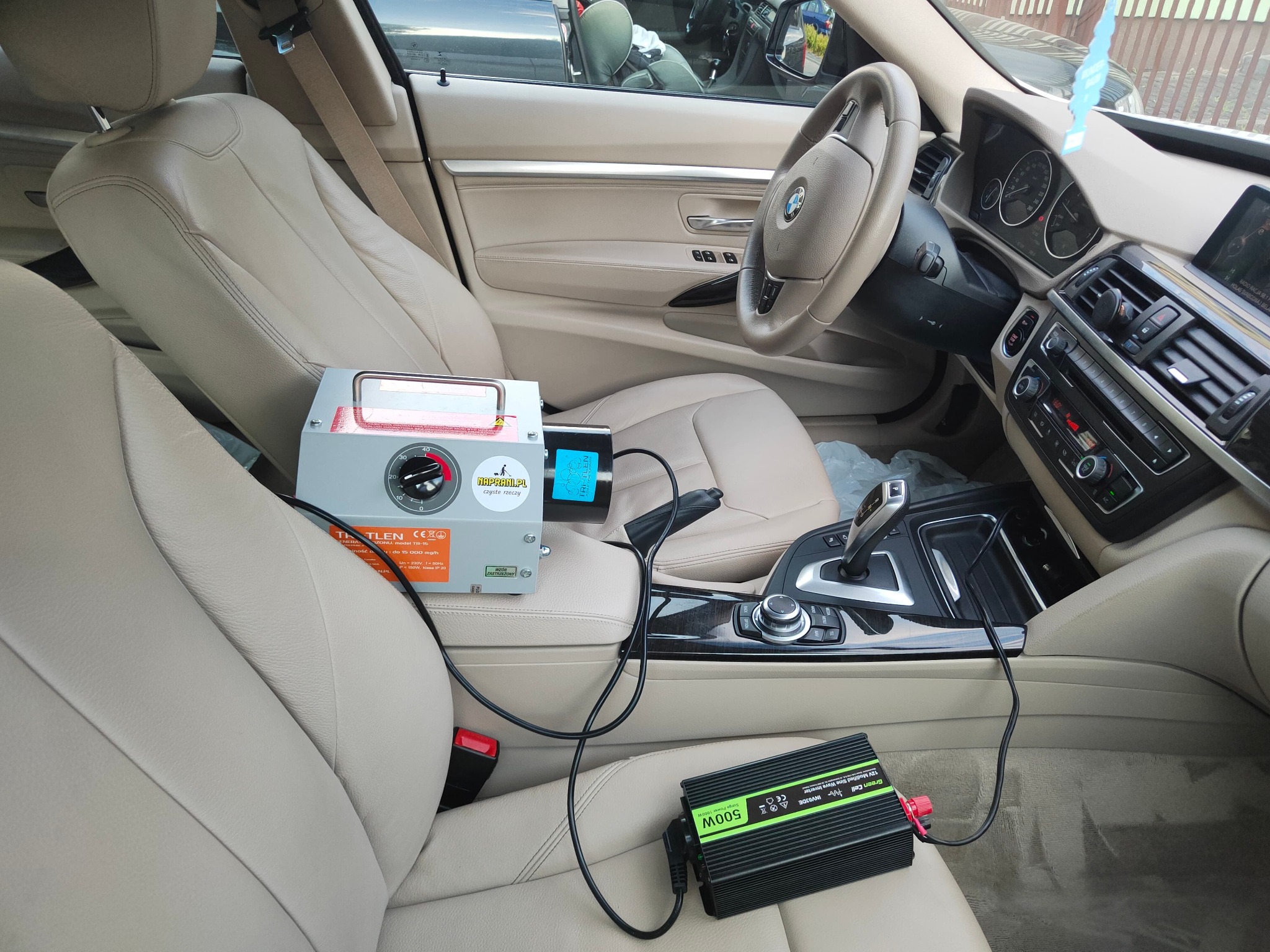 Wnętrze samochodu BMW z beżową tapicerką, na siedzeniu pasażera ustawione urządzenie do ozonowania powietrza marki Tri-Tlen oraz przetwornica napięcia Green Cell 500W, widoczne kable i elementy...