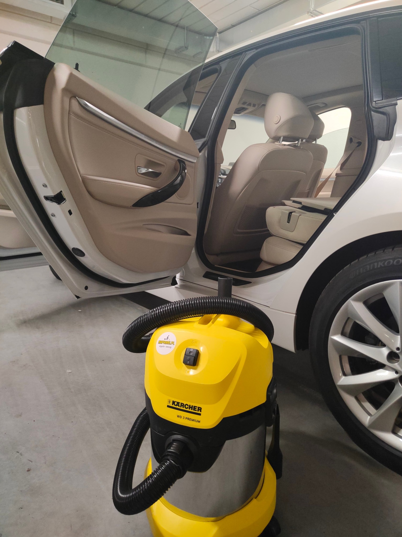 Żółty odkurzacz Kärcher WD 3 Premium stoi przed otwartymi drzwiami białego samochodu z beżową tapicerką.