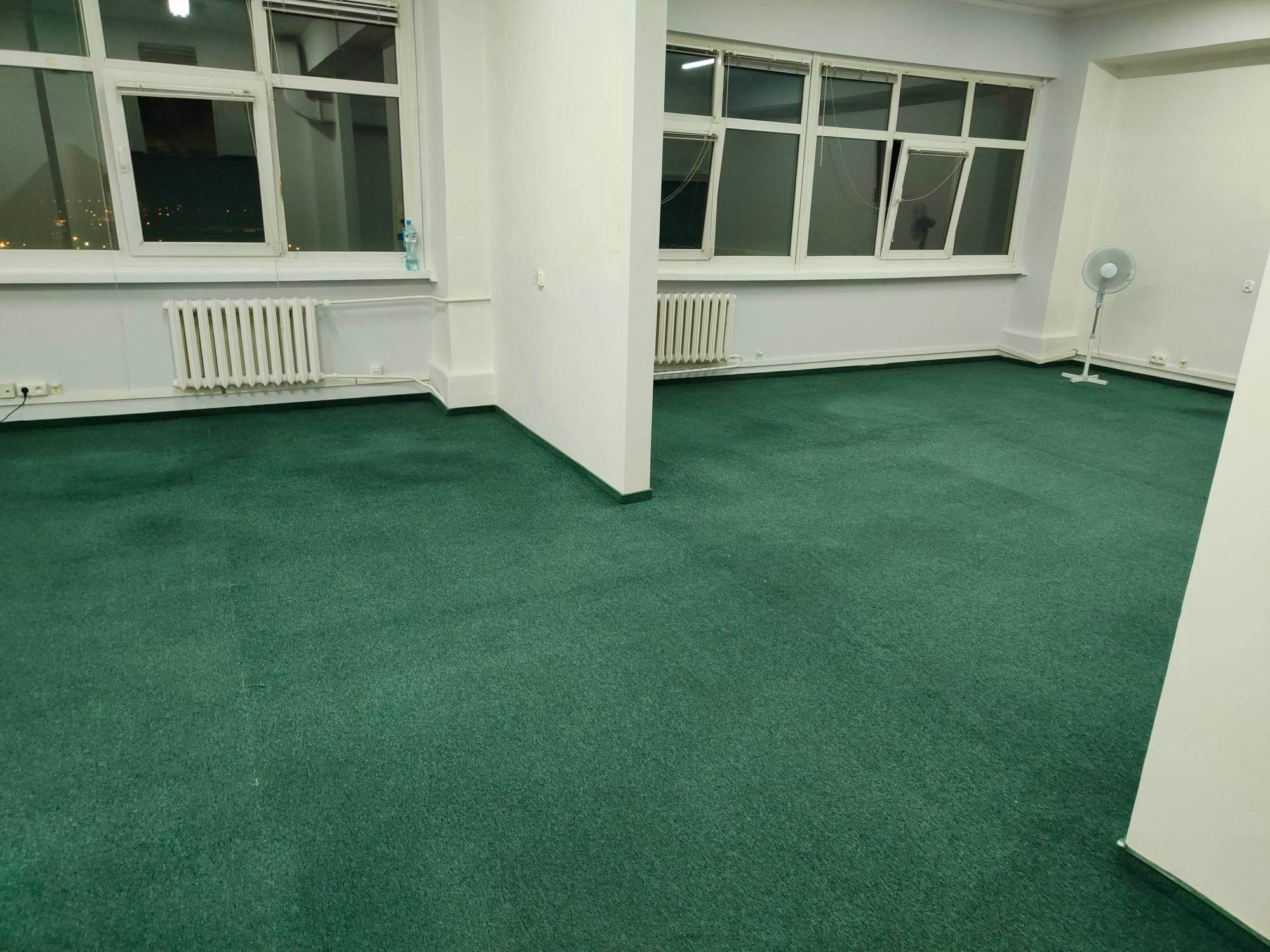 Puste pomieszczenie biurowe z zieloną wykładziną dywanową, białymi ścianami i grzejnikami pod oknami. W tle widoczny wentylator stojący.