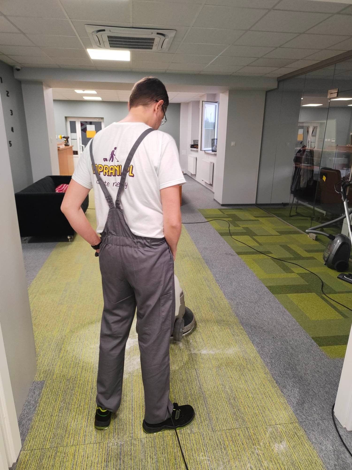 Pracownik w szarym kombinezonie czyści wykładzinę dywanową w biurze za pomocą profesjonalnej maszyny czyszczącej. Na plecach ma koszulkę z logo firmy sprzątającej.