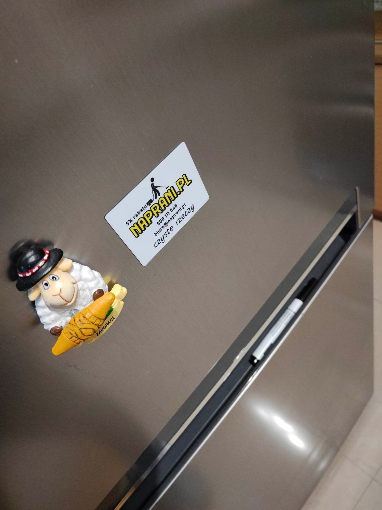 Lodówka ze stali nierdzewnej z magnesem owcy w kapeluszu trzymającej oscypka z Zakopanego i wizytówką firmy Naprani.pl oferującej czyste rzeczy.