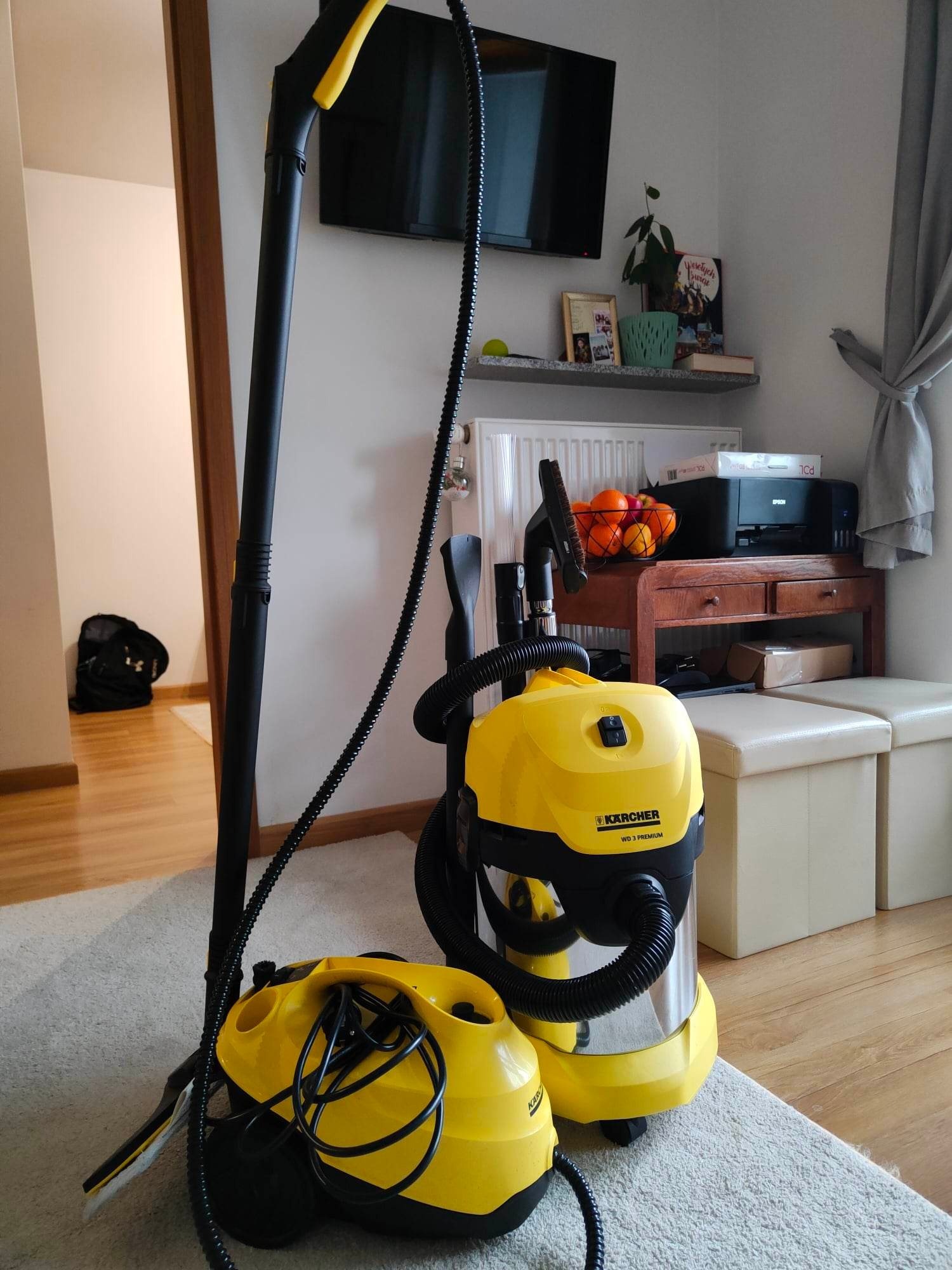 Dwa żółte odkurzacze Karcher WD 3 Premium i parownica na dywanie w mieszkaniu, widoczny telewizor i półka z dekoracjami w tle.