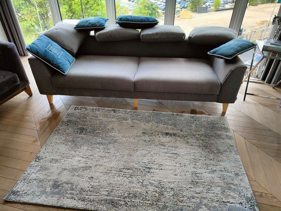 Szary, dwuosobowy tapicerowany fotel z drewnianymi nóżkami, ozdobiony niebieskimi poduszkami z białym wzorem, na tle wzorzystego dywanu w odcieniach szarości i beżu oraz parkietu ułożonego w jodełkę.