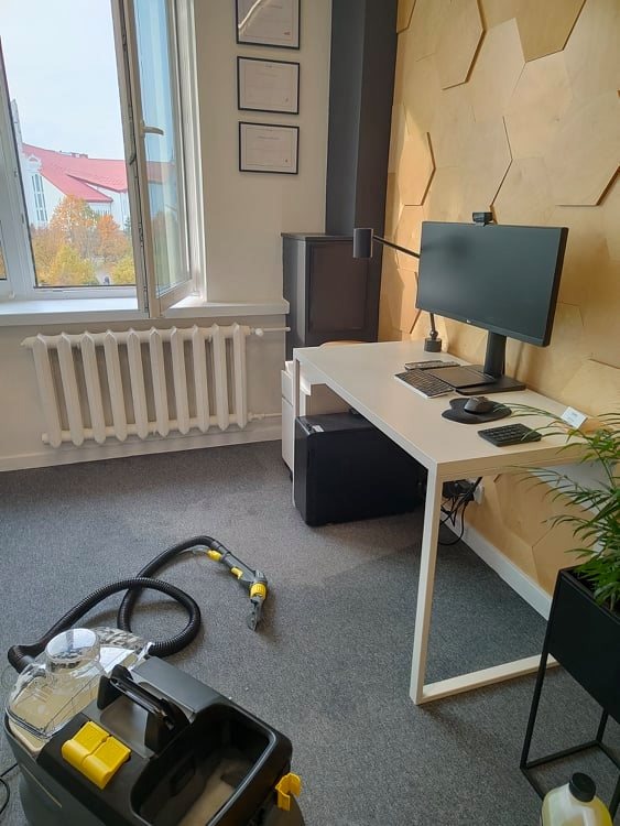 Profesjonalny odkurzacz piorący na szarym dywanie w biurze, widoczny fragment biurka i okno z widokiem na budynek z czerwoną dachówką.