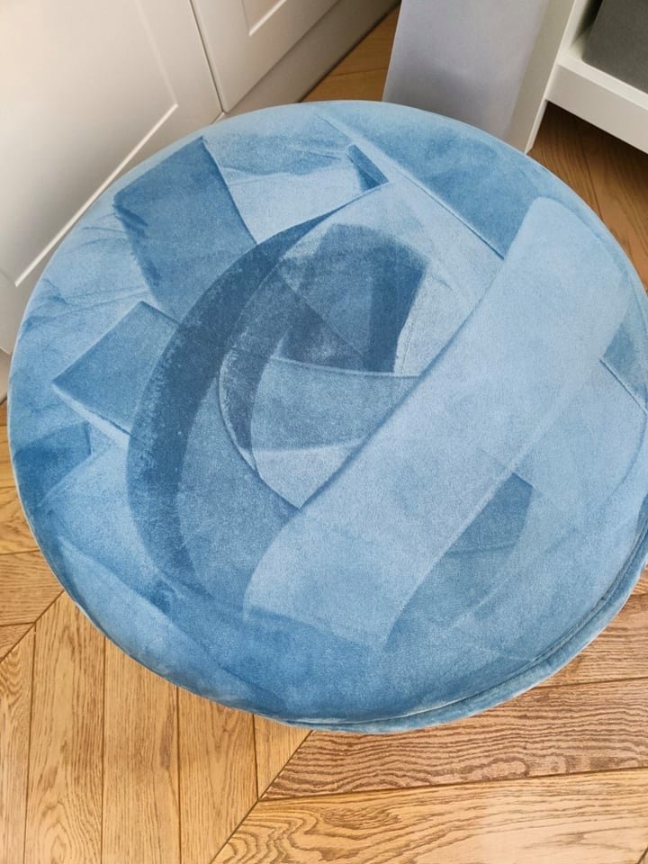 Okrągły, niebieski puf tapicerowany z abstrakcyjnym wzorem w jaśniejszych odcieniach niebieskiego, stojący na parkiecie.