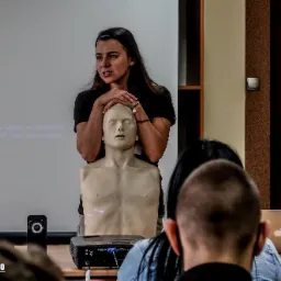 Instruktorka z długimi włosami demonstruje ułożenie rąk na manekinie podczas kursu pierwszej pomocy, w tle widoczny projektor i slajd prezentacji.