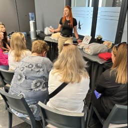 LIVE AND RESCUE - Instruktorka z rudymi włosami demonstruje na manekinie zasady udzielania pierwszej pomocy grupie osób siedzących na krzesłach.