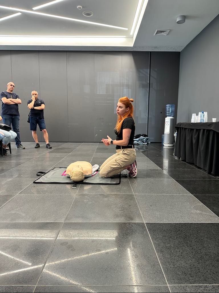 Instruktorka z rudymi włosami na kolanach demonstruje resuscytację na manekinie przed grupą obserwatorów w nowoczesnym wnętrzu z szarą podłogą i ścianami.