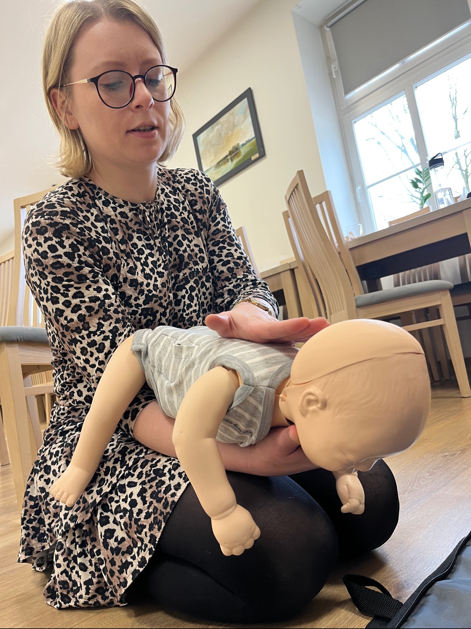 Instruktorka w okularach demonstruje ułożenie dłoni na klatce piersiowej manekina niemowlęcia podczas ćwiczeń resuscytacji krążeniowo-oddechowej.