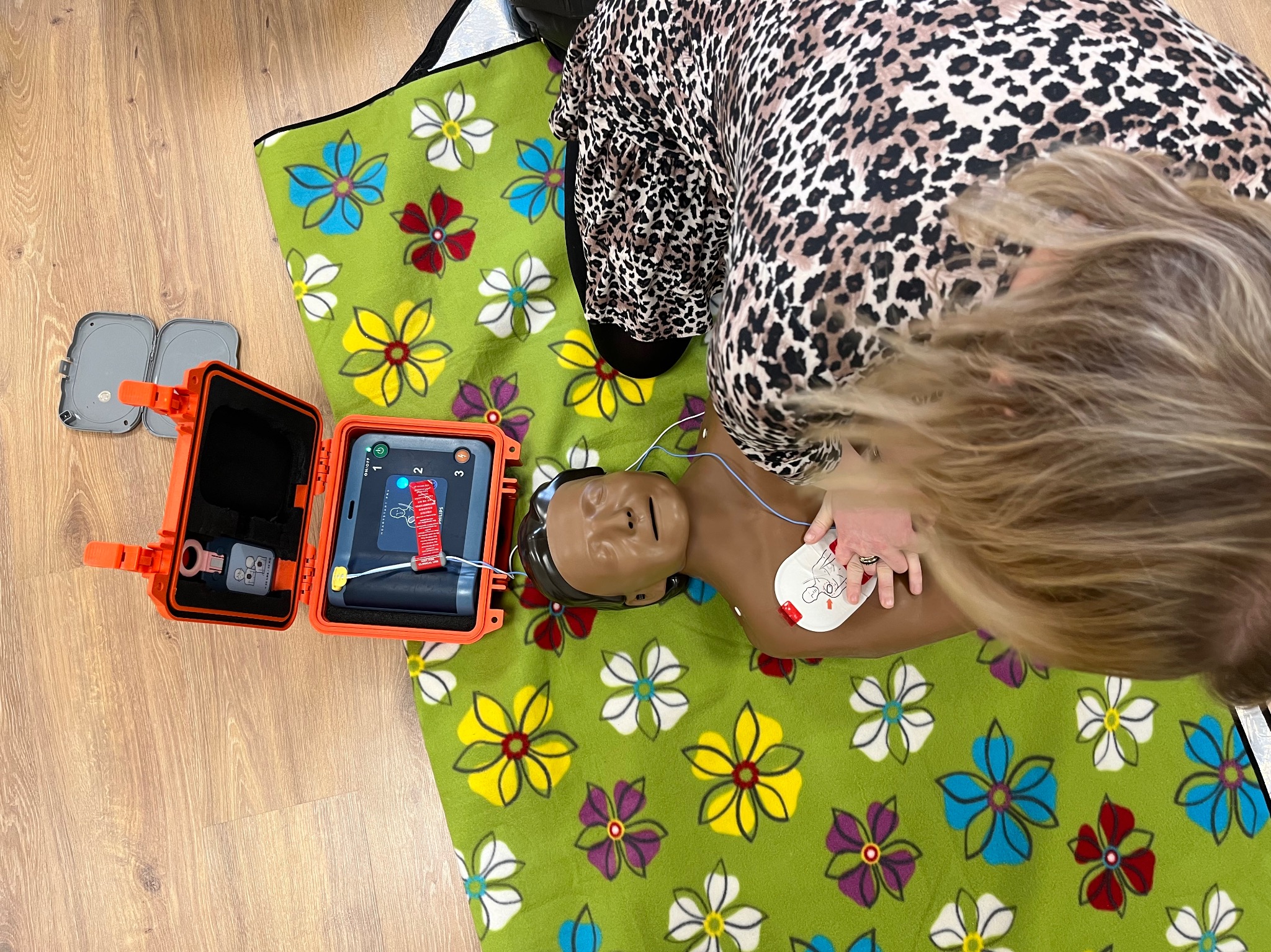 Instruktorka w bluzce w panterkę demonstruje użycie defibrylatora AED na manekinie leżącym na zielonym kocu w kwiaty, z otwartym pomarańczowym pudełkiem AED obok.