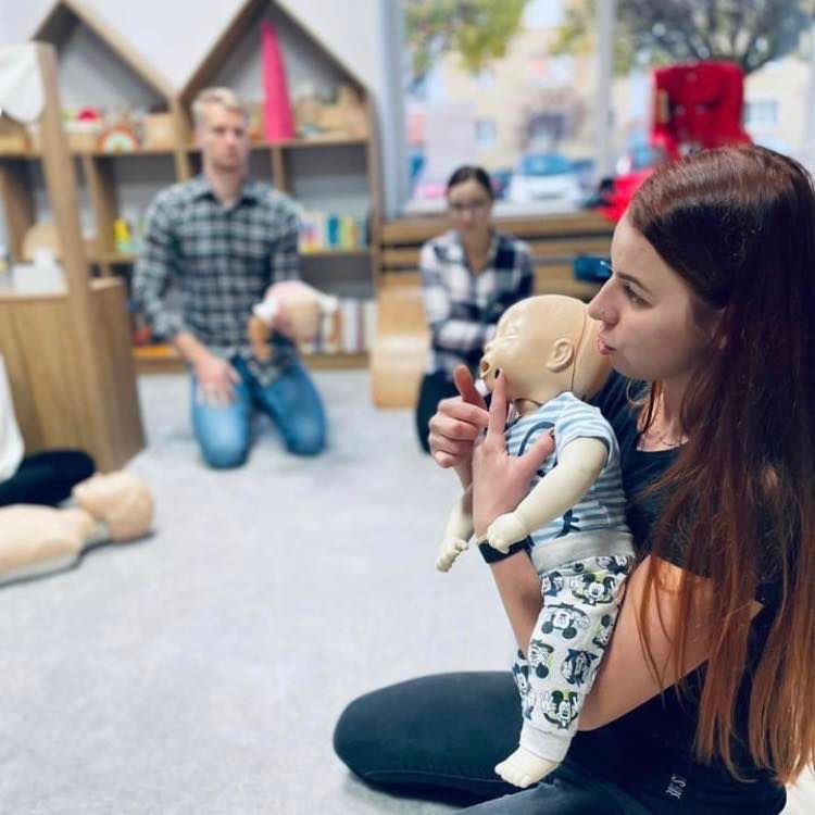 Instruktorka demonstruje technikę udrażniania dróg oddechowych niemowlęcia na manekinie podczas zajęć, w tle uczestnicy kursu i regały z książkami.