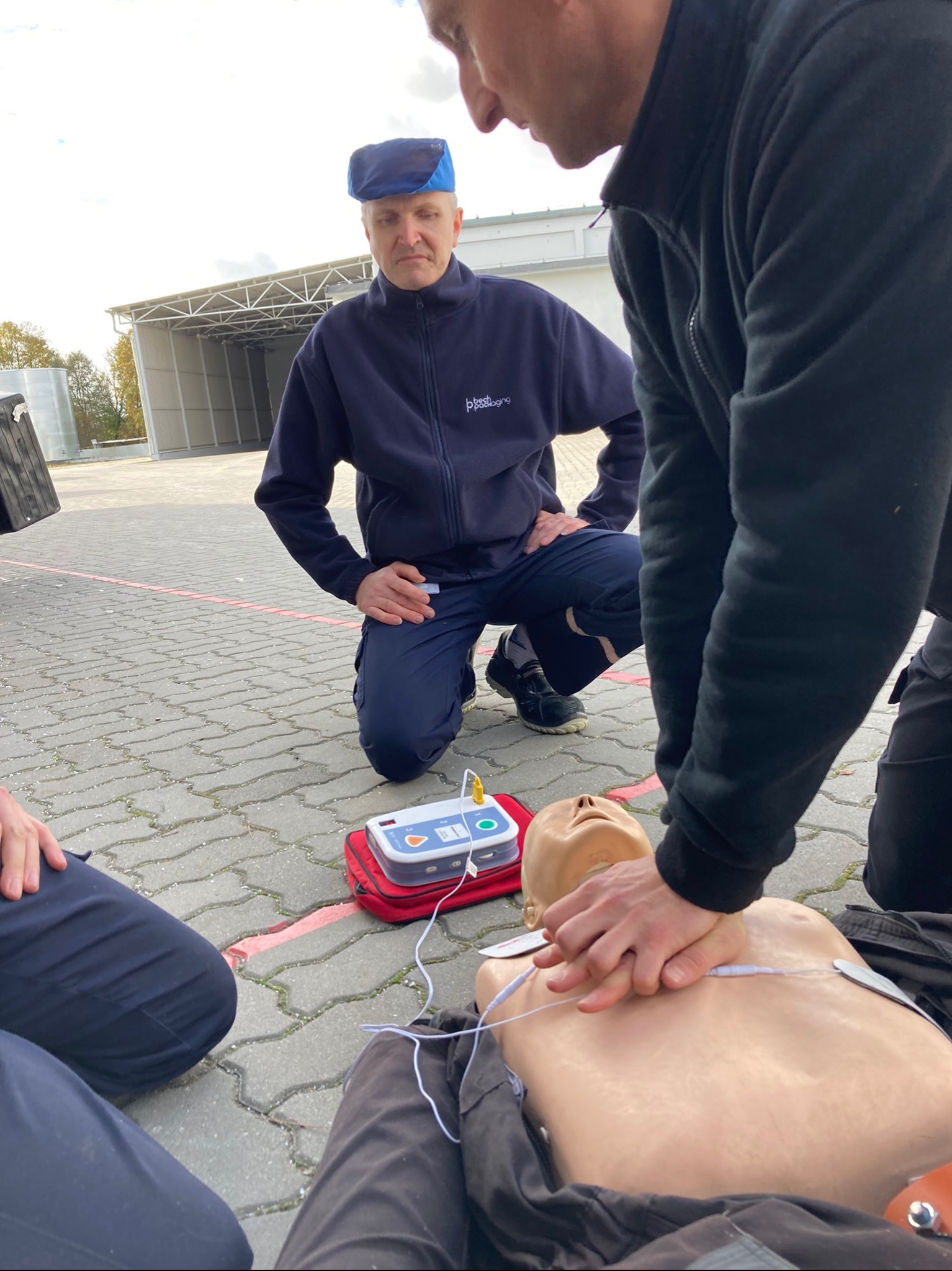 Instruktor demonstruje resuscytację krążeniowo-oddechową na manekinie, z użyciem defibrylatora AED na czerwonej torbie, podczas szkolenia na zewnątrz.