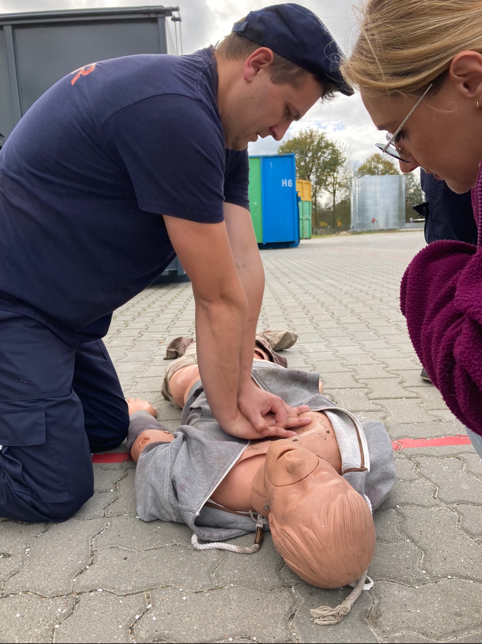 Instruktor demonstruje uciskanie klatki piersiowej na manekinie podczas praktycznego szkolenia, z bliska obserwuje kobieta w okularach.