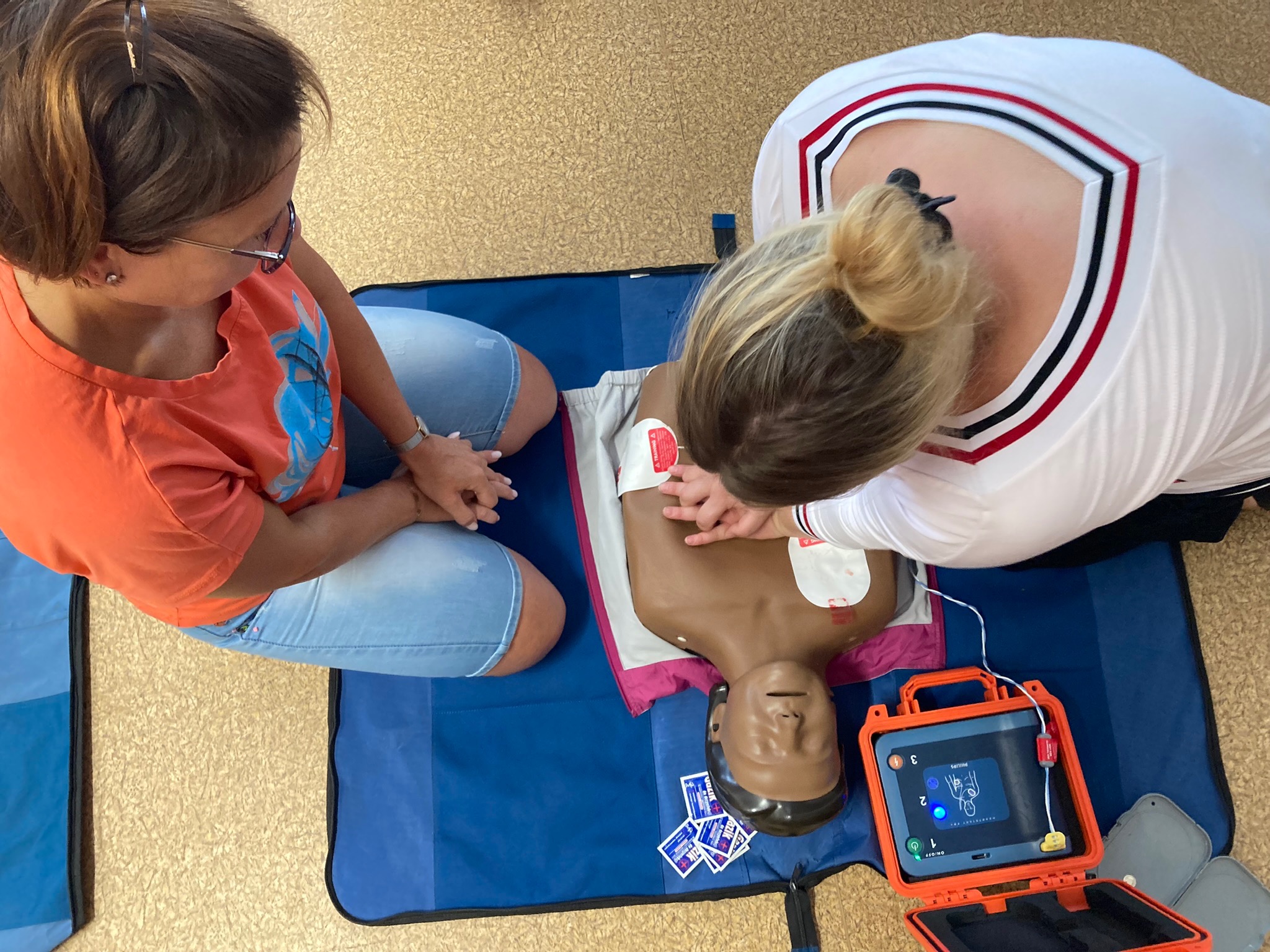 Instruktorzy demonstrujący resuscytację krążeniowo-oddechową na manekinie z użyciem defibrylatora AED w pomarańczowej obudowie.