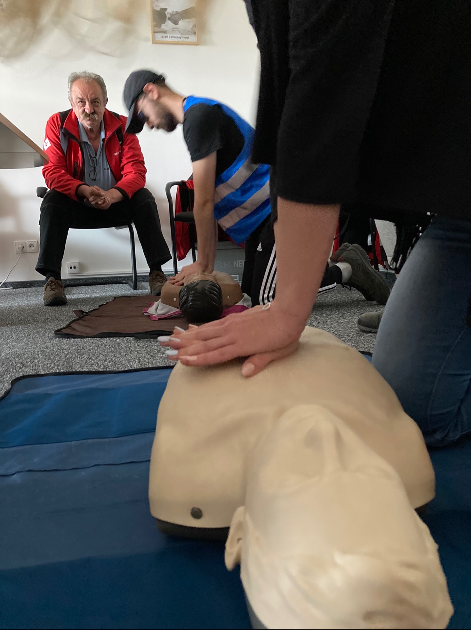 Instruktorzy demonstrują resuscytację krążeniowo-oddechową na manekinie podczas kursu pierwszej pomocy; widoczny starszy mężczyzna w czerwonej kurtce obserwujący ćwiczenie.