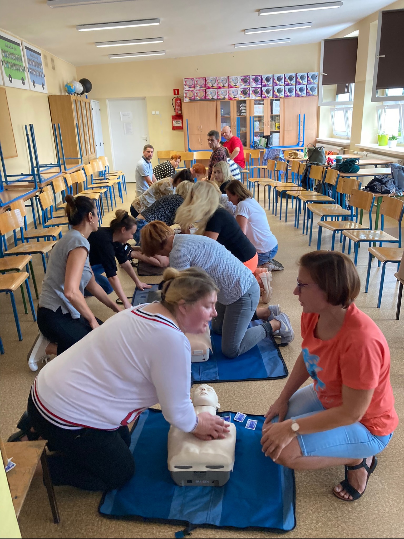 Grupa osób ćwiczy resuscytację krążeniowo-oddechową na manekinach w sali z krzesłami i szafkami, część osób obserwuje.