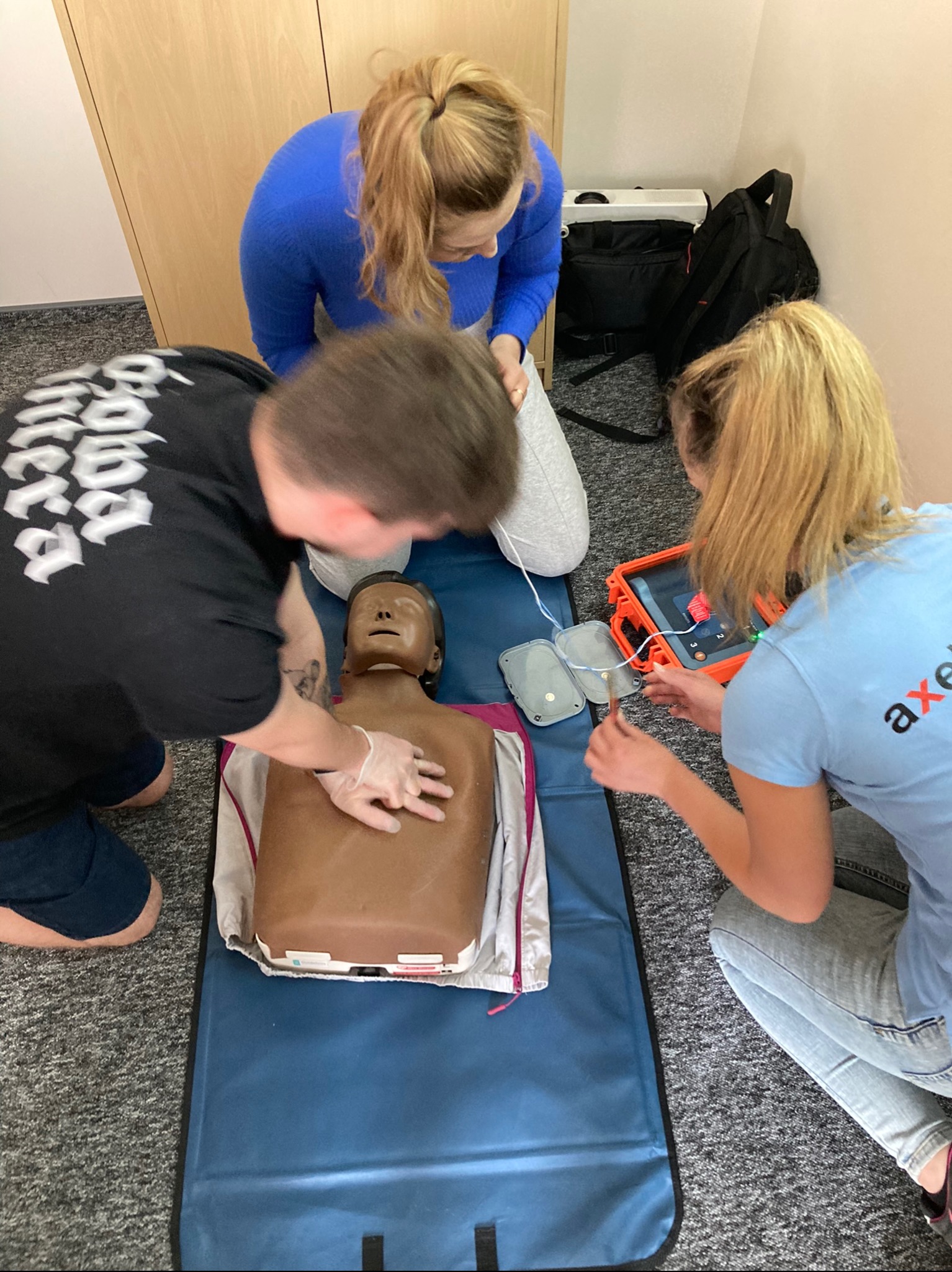 Trzy osoby ćwiczą resuscytację krążeniowo-oddechową na fantomie z użyciem defibrylatora, widoczne elektrody i pomarańczowa walizka defibrylatora.