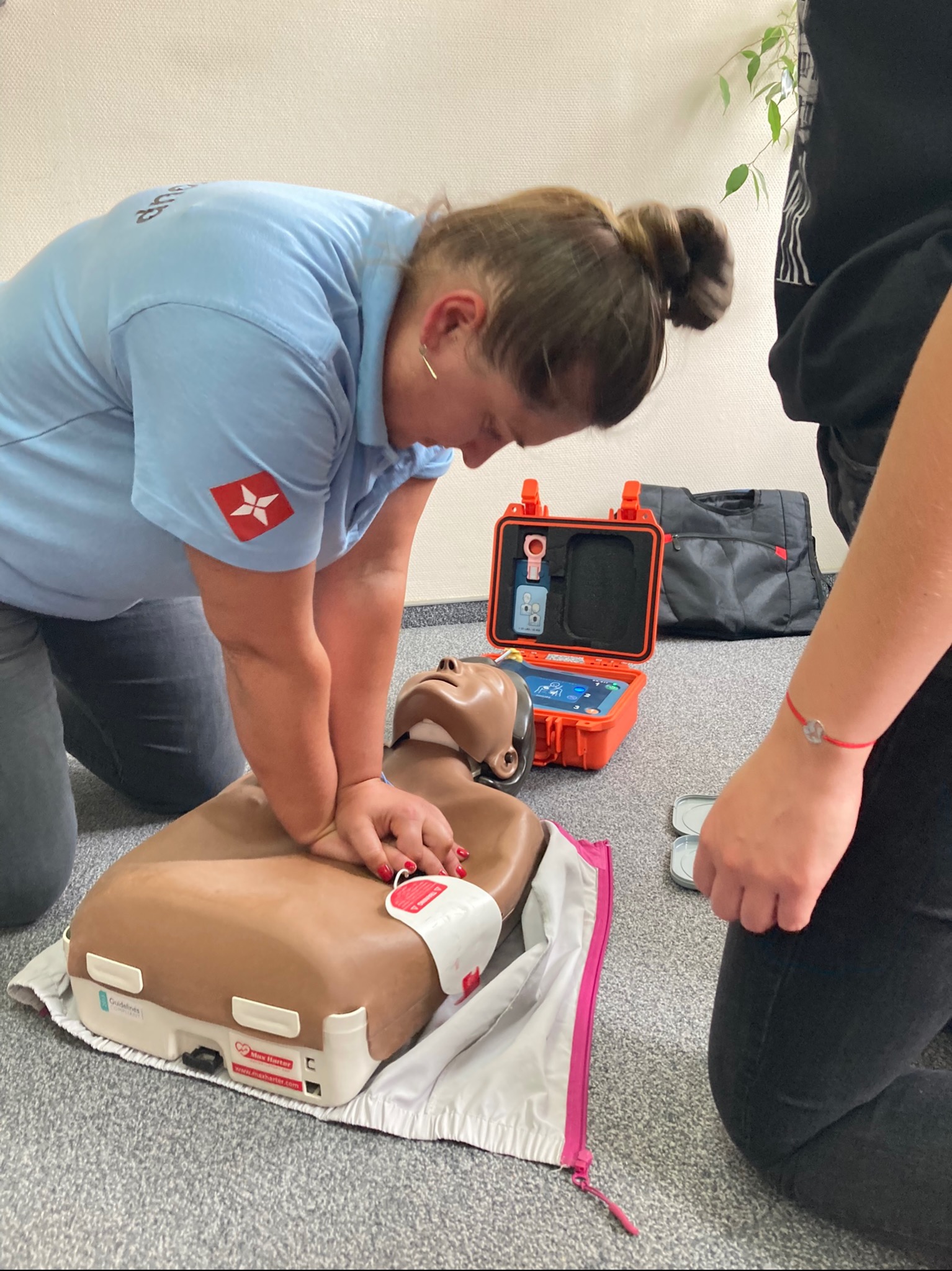 Instruktorka w błękitnej koszulce z logo, demonstruje uciskanie klatki piersiowej na manekinie do resuscytacji krążeniowo-oddechowej, obok otwarte pomarańczowe pudełko z urządzeniem treningowym.