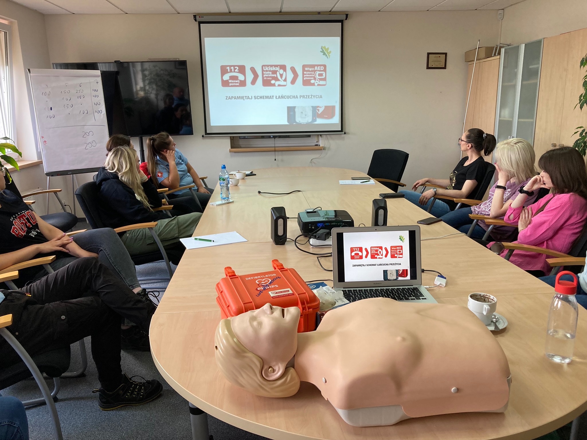 Sala szkoleniowa z manekinem do resuscytacji, defibrylatorem i laptopem wyświetlającym schemat postępowania, uczestnicy siedzą przy stole