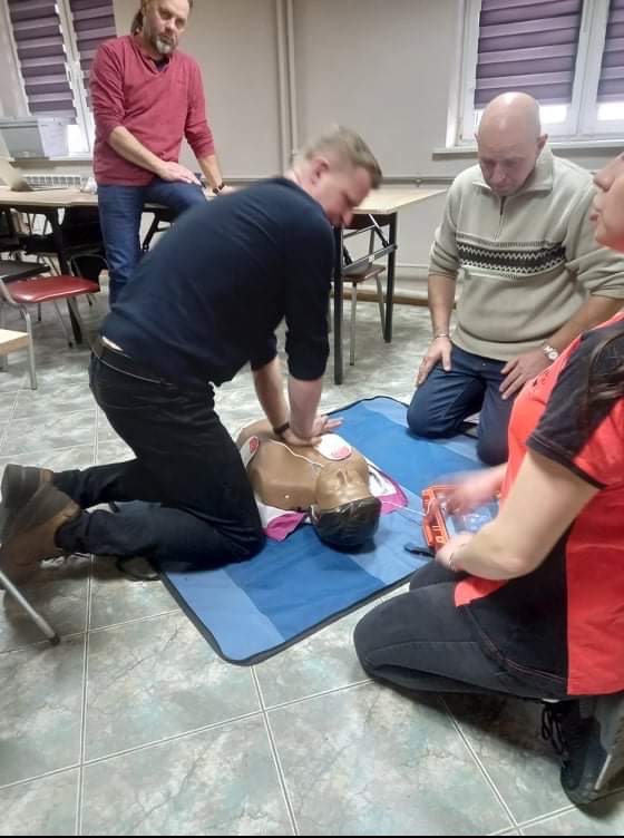 Instruktor demonstruje resuscytację krążeniowo-oddechową na manekinie podczas zajęć grupowych, z widoczną apteczką i uczestnikami obserwującymi.