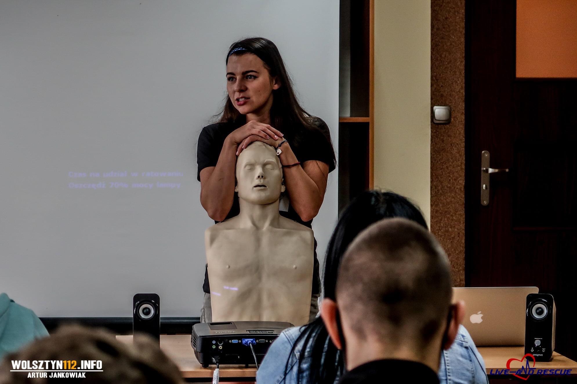 Instruktorka z długimi włosami demonstruje ułożenie rąk na manekinie podczas kursu pierwszej pomocy, w tle widoczny projektor i slajd prezentacji.