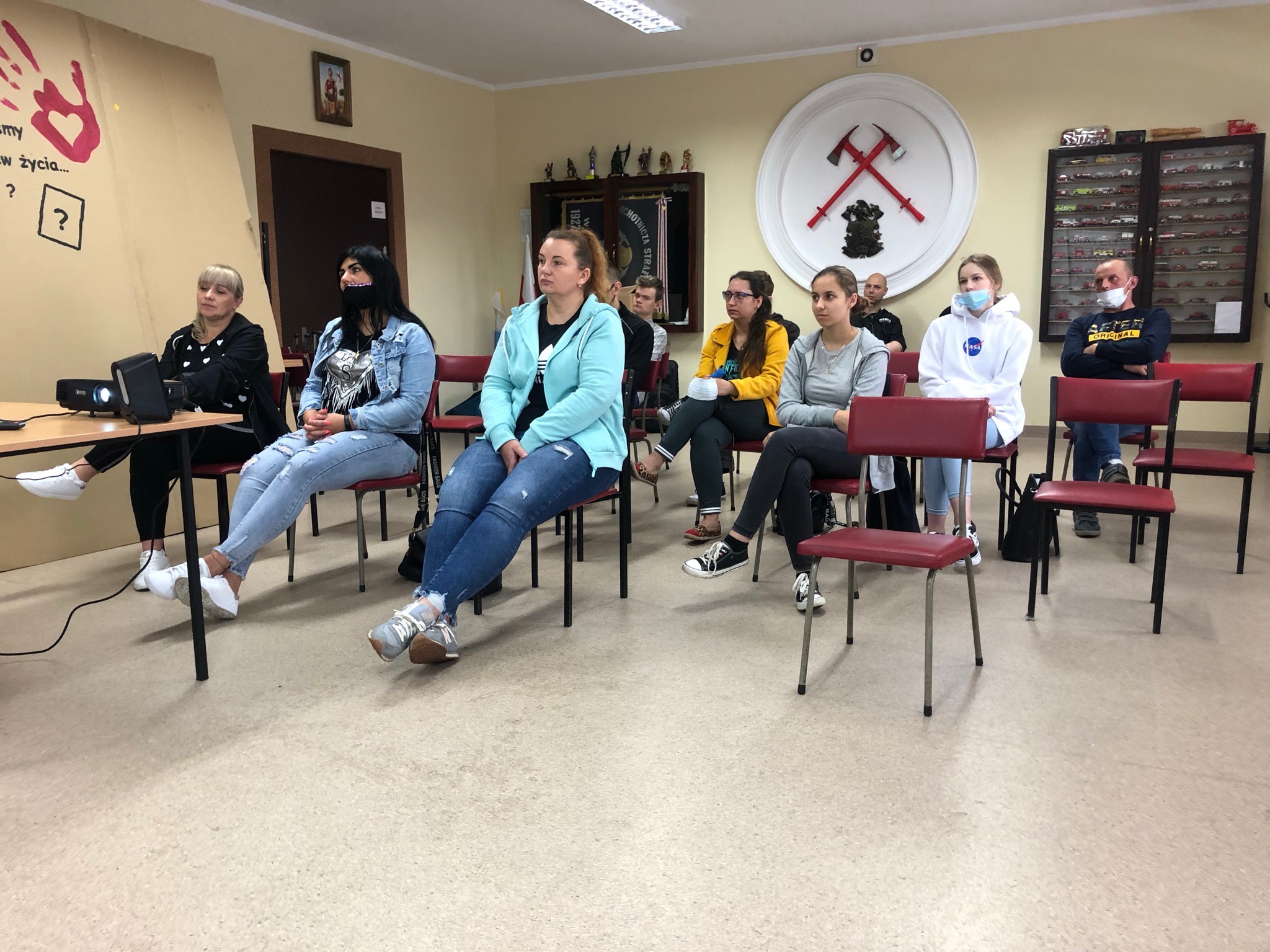 Grupa osób siedzi na krzesłach w sali, oglądając prezentację wyświetlaną z projektora. Na ścianie widoczny plakat z rysunkiem dłoni i pytajnikiem oraz logo ochotniczej straży pożarnej.