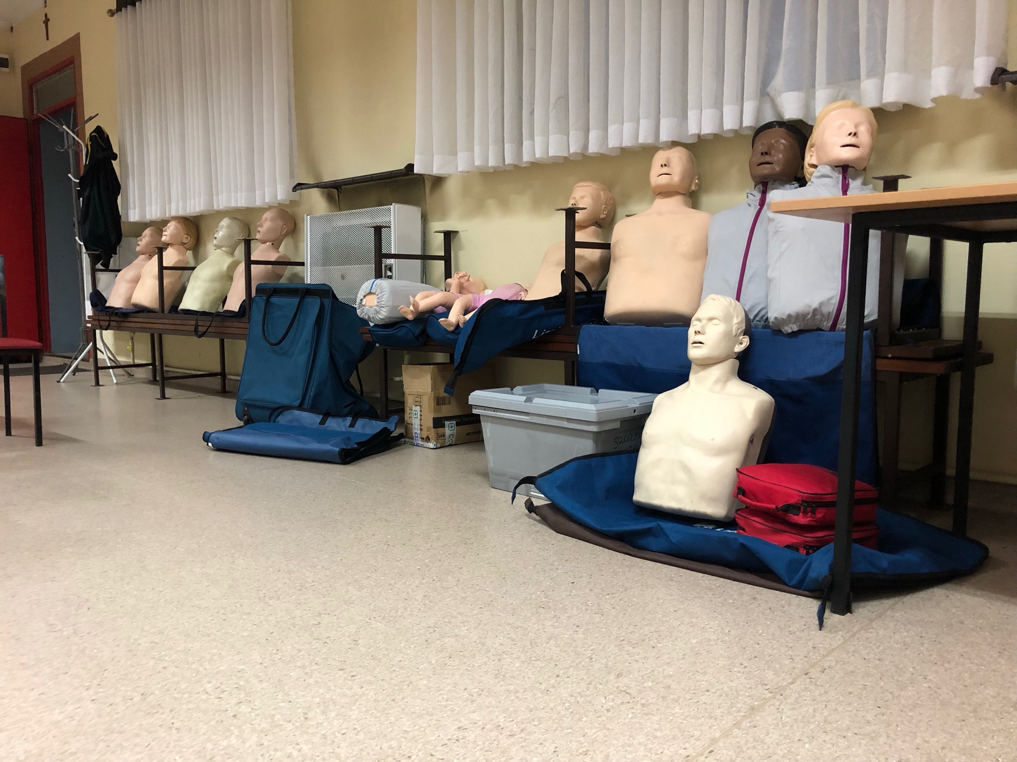 Sala szkoleniowa z manekinami do ćwiczeń resuscytacji, w tle białe zasłony i grzejnik.