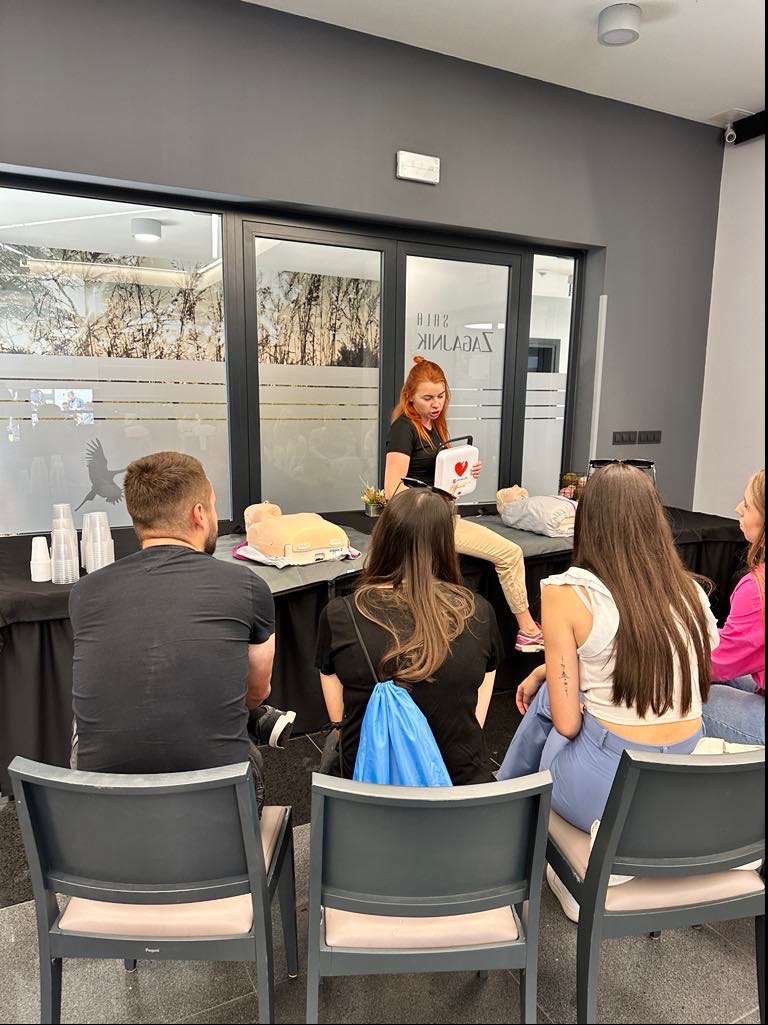 Instruktorka z czerwoną czupryną demonstruje użycie defibrylatora AED na tle sali z uczestnikami szkolenia i fantomami do ćwiczeń.