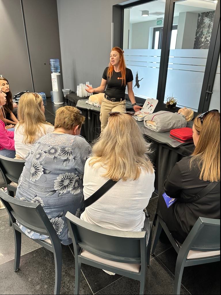 Instruktorka z rudymi włosami demonstruje na manekinie zasady udzielania pierwszej pomocy grupie osób siedzących na krzesłach.