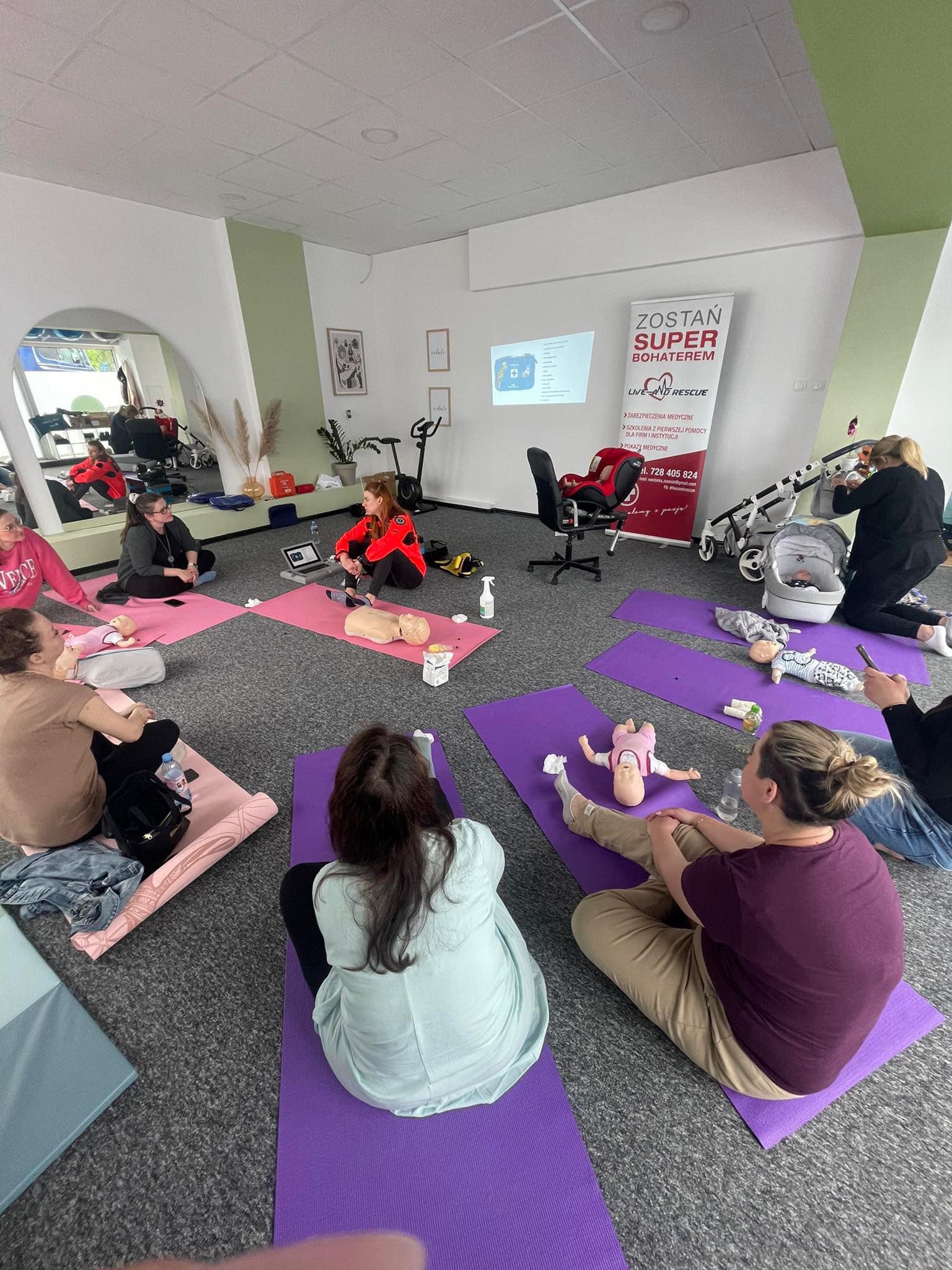 Grupa osób siedzi na matach podczas szkolenia z pierwszej pomocy, manekiny niemowląt i osoby dorosłe służą do ćwiczeń, na ścianie wyświetlany jest plakat informacyjny.
