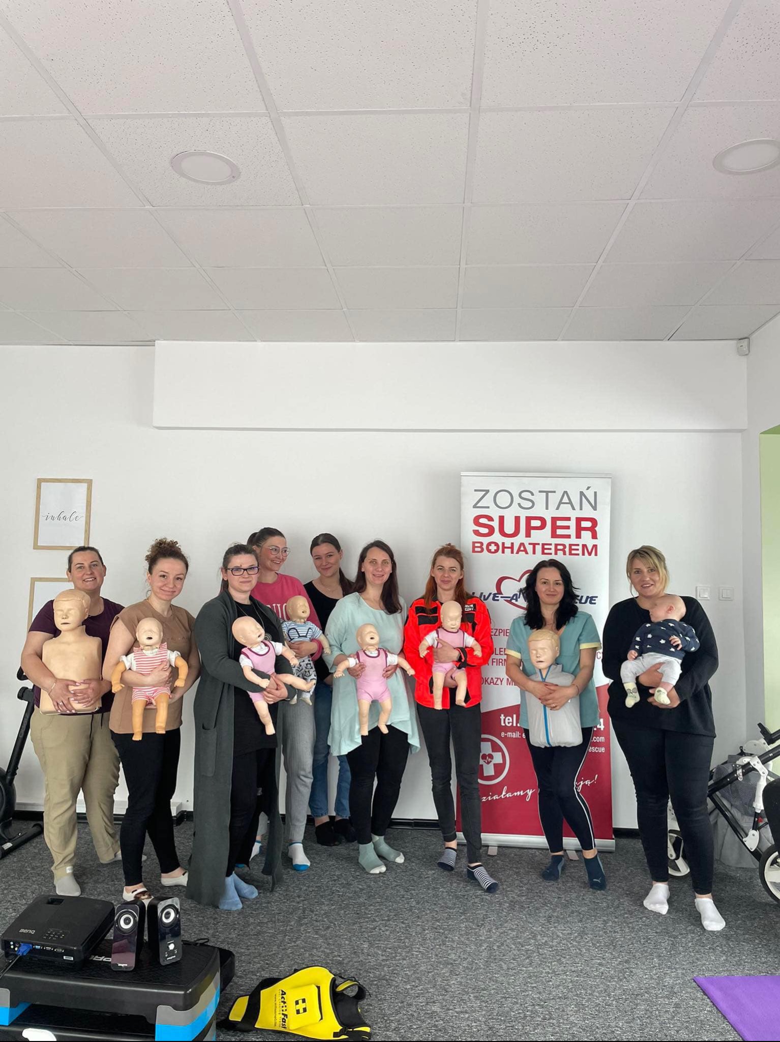 Grupa kobiet uczestniczy w kursie pierwszej pomocy, trzymając manekiny niemowląt i dzieci, z banerem 'Zostań Super Bohaterem' w tle, w jasnym pomieszczeniu.