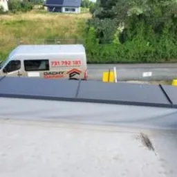 Widok z góry na fragment płaskiego dachu pokrytego szarą membraną, z widocznym samochodem dostawczym firmy dekarskiej w tle i zielenią otaczającą budynek.