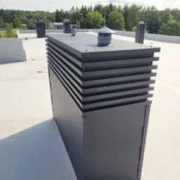 Szary, metalowy komin wentylacyjny na płaskim dachu pokrytym jasną membraną. W tle widoczny las i fragment budynku.