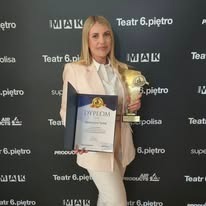 Blond kobieta w beżowym żakiecie trzyma dyplom i statuetkę na tle ściany z logo Teatru 6. piętro i MAK. Uśmiech, frontowe ujęcie, dobre oświetlenie.