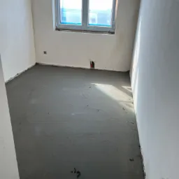 Świeżo wylana, szara posadzka w surowym pomieszczeniu z oknem; widoczne ślady po pracy i niedoskonałości powierzchni.