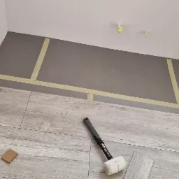 Proces układania paneli podłogowych w rogu pomieszczenia, widoczna gumowa młotek, przycięte panele i kątownik, ściany zabezpieczone taśmą malarską.
