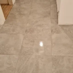 Ułożona jasnoszara podłoga z płytek ceramicznych w trakcie remontu, widoczne niedokończone obramowanie drzwi i wystające kable ze ściany.