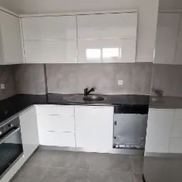 Nowoczesna kuchnia z białymi szafkami, czarnym blatem i szarymi płytkami na ścianie, widoczny okap, zlew i częściowo lodówka.