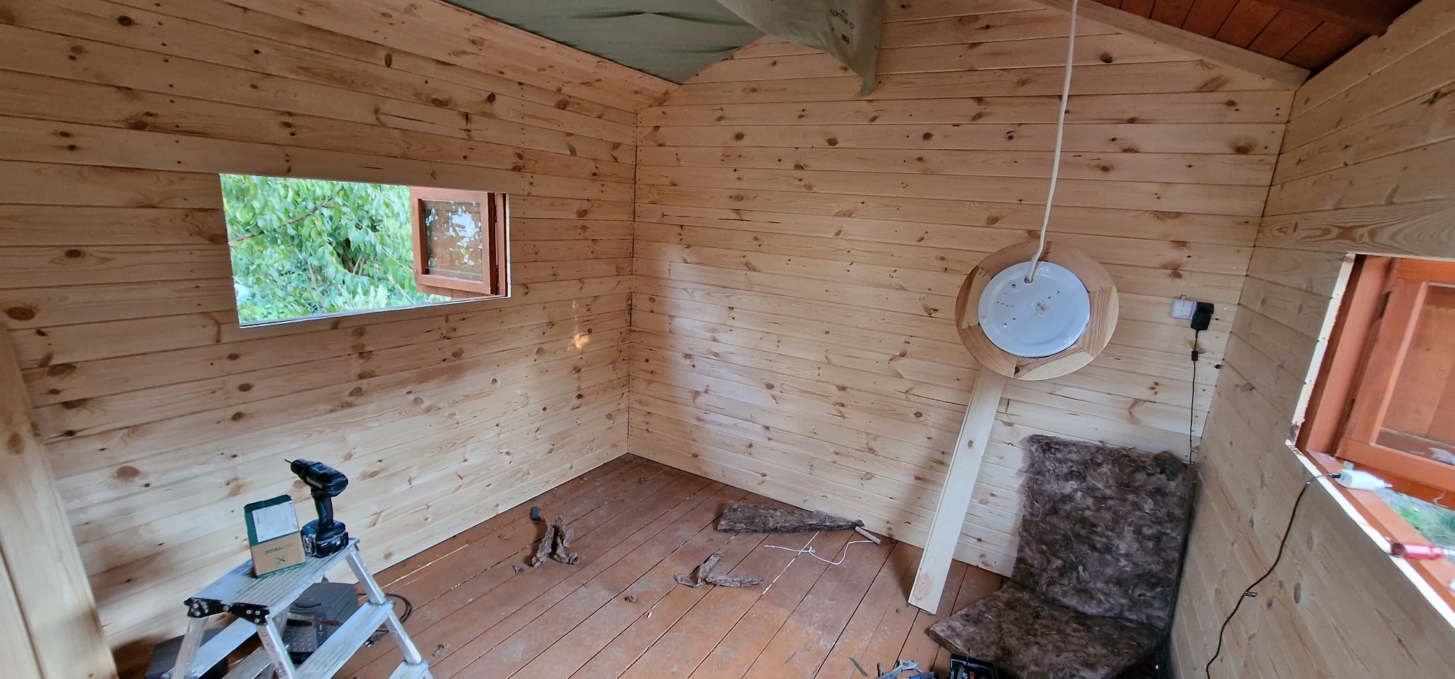 Wnętrze drewnianej konstrukcji w trakcie wykańczania, widoczne deski na ścianach i podłodze, okno z widokiem na zieleń, drabina z wiertarką, fragment ocieplenia oraz prowizoryczne oświetlenie...